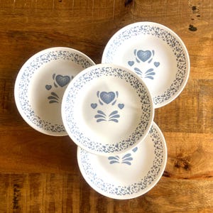 May include: Four white ceramic plates with a blue heart and floral design. The plates have a speckled blue border and are arranged on a wooden surface. The plates are a vintage style.