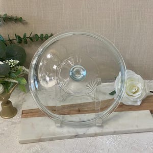 May include: A circular, clear glass lid featuring a raised centre knob surrounded by a sunburst pattern of fine, radiating lines. The lid has a smooth, slightly domed surface and a thick, rounded outer rim designed for a secure fit on cookware.