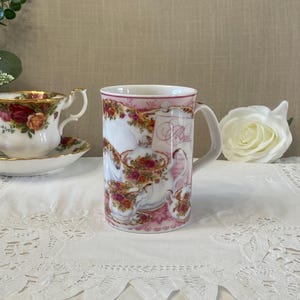 Peut inclure: Une tasse en céramique blanche avec un motif rose et floral. La tasse présente une anse et un motif décoratif de tasses à thé, de théières et de roses. Le mot "Menu" est visible sur la tasse. Une tasse et une soucoupe sont visibles en arrière-plan.