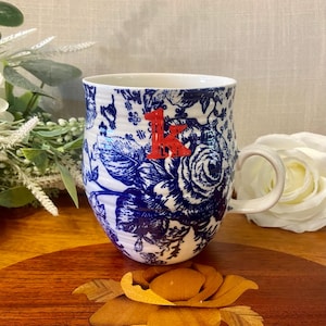 May include: A white ceramic mug featuring a blue floral pattern and a prominent red letter K printed on the front. The mug has a rounded body, a curved handle, and a textured surface with subtle horizontal ridges throughout the exterior design.