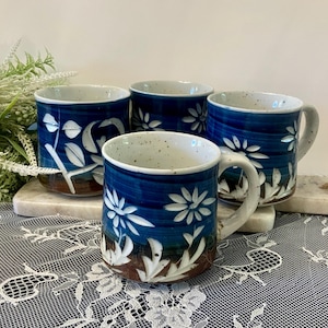 May include: Four ceramic mugs with a dark blue glaze and white floral designs. The mugs have a speckled white interior and brown base. The mugs are arranged on a white marble tray.