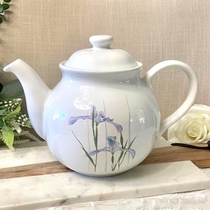May include: A white ceramic teapot featuring a rounded body, a curved spout, and a matching lid. The surface is decorated with a delicate illustration of purple iris flowers with green stems and faint vertical pastel stripes on the front.