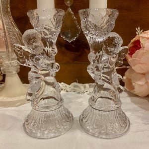 May include: Two clear glass candle holders, each with an angel figurine. The candle holders have a detailed design and hold white candles. The background includes a decorative glass piece and a pink flower arrangement.