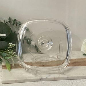 May include: A clear, square glass lid with a central knob. The lid is slightly rounded and has a transparent appearance. It is displayed on a clear stand, suggesting it's a kitchen or dining item. The background includes greenery and a marble-like surface.