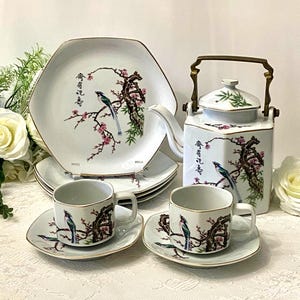 May include: A white porcelain tea set featuring a bird and floral design. The set includes a teapot, cups, saucers, and plates, all with gold trim. The design features a bird perched on a flowering branch. The teapot has a brass handle.