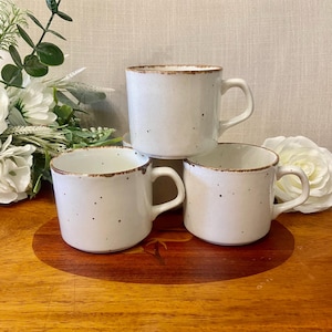 May include: A set of three off-white ceramic mugs with a speckled pattern and a brown rim. Each mug features a cylindrical body and a curved handle. The mugs are arranged in a cluster, with one mug positioned behind the other two in the front.