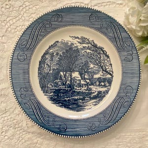 May include: Blue and white decorative plate with a rural landscape design. The plate features a detailed illustration of a countryside scene with trees, a river, and buildings. The rim has a textured pattern and decorative swirls.