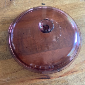 May include: A round, translucent purple glass lid featuring a smooth, domed surface and a central knob handle. The area surrounding the knob is decorated with a series of fine, radiating lines that extend outward, creating a subtle sunburst pattern.