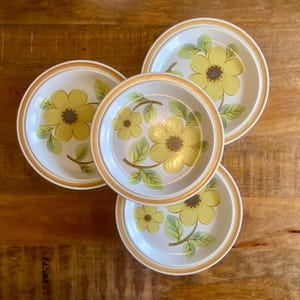 May include: Four white ceramic plates with a yellow floral design and a brown rim. The plates feature a large yellow flower with a brown centre and green leaves. The plates are arranged on a wooden surface.