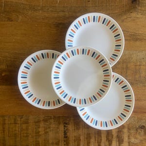 May include: Four white ceramic plates with a striped border in blue, orange, and beige. The plates are arranged on a wooden surface, showcasing a simple yet stylish design. Perfect for serving meals or as decorative pieces.