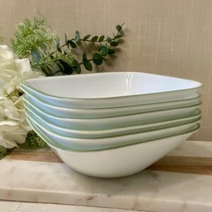 May include: A stack of six white square bowls with a light green trim. The bowls are stacked on top of each other. The bowls are sitting on a marble and wood cutting board.