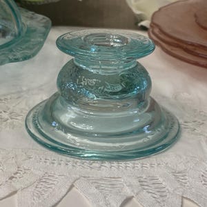 May include: A clear, light blue glass candlestick holder with a decorative floral pattern. The candlestick holder has a round base, a bulbous centre, and a flat top. The glass has a slight greenish tint. The candlestick holder is on a white lace tablecloth.