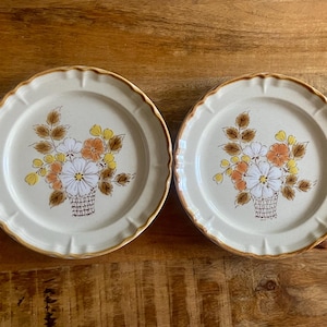 May include: Two round ceramic plates with a cream-coloured base and a scalloped edge featuring a brown rim. The centre of each plate displays a floral illustration of white, orange, and yellow flowers with brown leaves arranged in a basket.