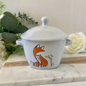 May include: A white ceramic lidded bowl with a speckled finish. The bowl features a cartoon fox illustration in orange and black with a small tree design. The bowl has two small handles and a round knob on the lid.
