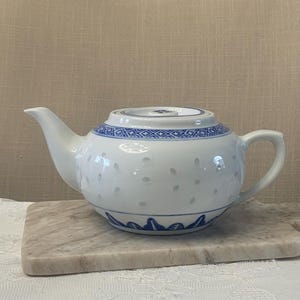 May include: White teapot with a blue and white floral design. The teapot has a lid and a spout. It is sitting on a marble surface.