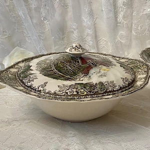 May include: Cream-coloured serving bowl with a lid, featuring a detailed landscape design in green, brown, and white. The rim and lid edge have a decorative pattern. A small knob sits atop the lid. Part of a matching set.