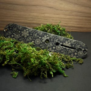 Könnte beinhalten: Ein schwarzes Metallwerkzeug mit Paisley-Muster, das kreisförmige und florale Designs aufweist. Das Werkzeug hat mehrere Löcher und einen Schlitz und liegt auf einem Bett aus grünem Moos. Der Hintergrund ist dunkles Holz.