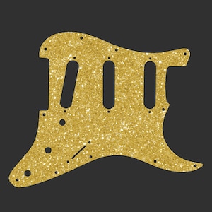 Puede incluir: Un golpeador de guitarra dorado con purpurina, con tres agujeros alargados y varios agujeros más pequeños para tornillos. El golpeador tiene una forma curva y está diseñado para adaptarse a un modelo de guitarra específico. El fondo es gris oscuro liso.