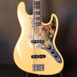 Puede incluir: Bajo eléctrico color crema con un golpeador decorativo con un diseño de paisaje. El bajo tiene cuatro cuerdas, pastillas negras y herrajes cromados. El cuerpo muestra signos de desgaste.