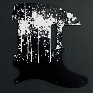 May include: Black guitar pickguard with a white splatter paint design. The pickguard has a unique shape with a cutout for the pickup and control knobs. The white paint is applied in a random pattern, creating a striking contrast against the black background.