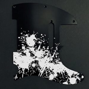 May include: Black guitar pickguard with a white splatter paint design. The pickguard has a unique shape with screw holes and cutouts for the guitar's components. The white paint splatters create a bold, artistic look.