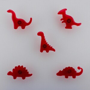 May include: Five red dinosaur-shaped buttons with a glossy finish on a white background. Each button features a different dinosaur design, including a Brontosaurus, Stegosaurus, and others. The buttons have a small, circular detail in the centre.