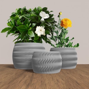 May include: Three grey ceramic planters with a wavy texture. The largest planter holds a green plant with white flowers. The middle planter is empty. The smallest planter holds a green plant with yellow flowers.