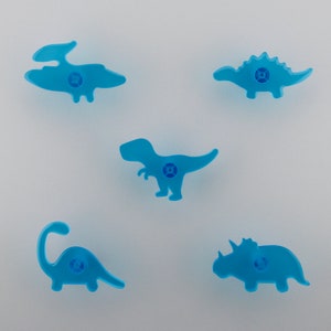 Peut inclure: Cinq boutons de meuble en forme de dinosaures bleu clair. Les boutons présentent différentes silhouettes de dinosaures, dont un T-Rex, un Stégosaure et un Tricératops. Chaque bouton a un trou de vis central pour une installation facile. Idéal pour une chambre d'enfant.