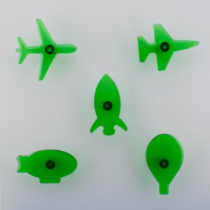 May include: Five translucent green transport-themed shapes: an aeroplane, a jet, a rocket, a blimp, and a hot air balloon. Each shape has a black circular detail in the centre, suggesting a fastener or decorative element. The objects are arranged against a white background.