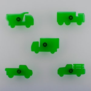 May include: Five green truck-shaped cabinet knobs. The knobs are in various truck designs, including a dumper truck, a flatbed truck, and a pickup truck. Each knob has a small, dark-coloured screw in the centre.