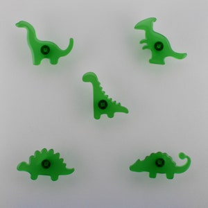 May include: Five green dinosaur-shaped cabinet knobs. Each knob features a different dinosaur design, including a Brontosaurus, Stegosaurus, and a T-Rex. The knobs are made of a translucent green material and have a screw in the centre.