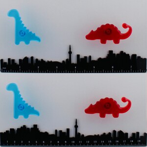 Op de afbeelding: Twee sets dinosaur-vormige kastknoppen in blauw en rood, met een zwarte skyline liniaal eronder. De linialen meten tot 48 cm. De knoppen zijn een leuke en kleurrijke toevoeging aan elk meubel.