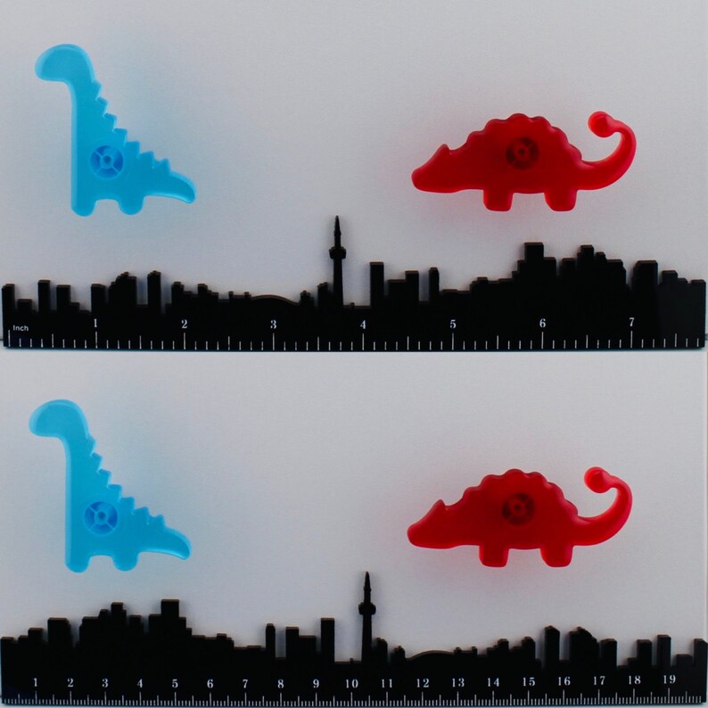 Op de afbeelding: Twee sets dinosaur-vormige kastknoppen in blauw en rood, met een zwarte skyline liniaal eronder. De linialen meten tot 48 cm. De knoppen zijn een leuke en kleurrijke toevoeging aan elk meubel.