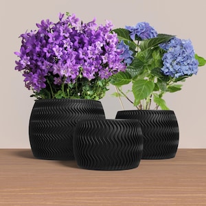 May include: Three black wavy textured ceramic pots with purple and blue flowers. The pots are different sizes and are arranged on a wooden surface.