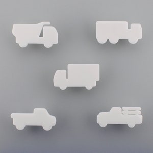 May include: Five white truck-shaped cutouts in various styles, including a dumper truck, a lorry, a box truck, and two pickup trucks. The cutouts are arranged against a grey background.