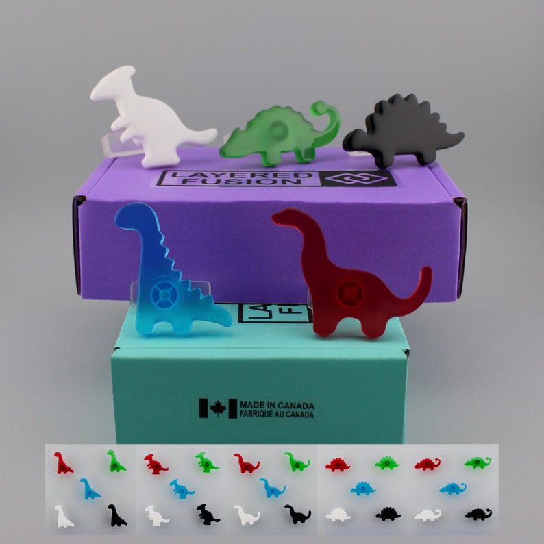Op de afbeelding: Een verzameling kleurrijke, dinosaurusvormige acrylspelden. De spelden zijn in verschillende kleuren, waaronder wit, groen, zwart, blauw en rood. De spelden worden weergegeven bovenop twee dozen, een paarse en een turquoise. De dozen hebben de tekst "LAYERED FUSION" en "MADE IN CANADA".