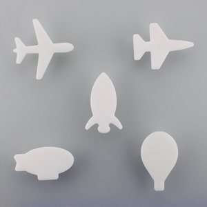 May include: Five white, stylized transport shapes are arranged on a grey background. The shapes include two aeroplanes, a rocket, a blimp, and a hot air balloon. The objects are likely decorative or craft elements.