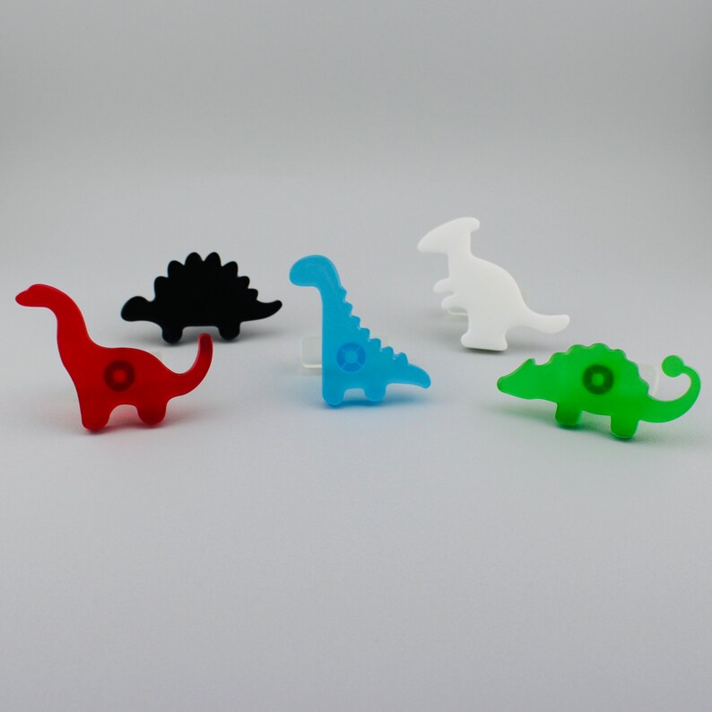 Op de afbeelding: Een verzameling van vijf kleurrijke dinosaurusknoppen. De knoppen zijn rood, zwart, blauw, wit en groen. Elke knop heeft een cirkelvormig ontwerp in het midden. Deze decoratieve knoppen zijn perfect voor een kinderkamer of een dinosaurusproject.