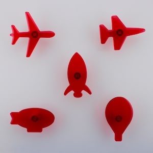 May include: Five red toy vehicles: an aeroplane, a jet, a rocket, a blimp, and a hot air balloon. Each toy has a circular design in the centre. The toys are arranged on a white background.