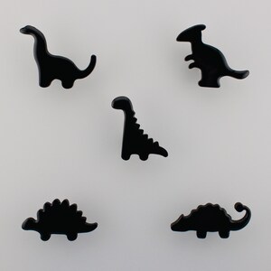 Peut inclure: Cinq éléments décoratifs noirs en forme de dinosaures. Les dinosaures sont dans diverses poses, dont un Brontosaure à long cou, un Stégosaure et un Parasaurolophus. Les accents sont brillants et sur fond blanc.