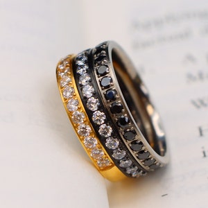 May include: Three stacked rings with a gold, silver, and black finish. Each ring is encrusted with small, evenly spaced gemstones. The rings are resting on the open pages of a book.