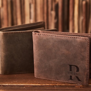 May include: Two brown leather wallets are displayed on a wooden surface. One wallet is open, revealing the interior, while the other is closed and features the name "RICHIE" with a stylized "R" initial. The wallets have a classic, rugged appearance.
