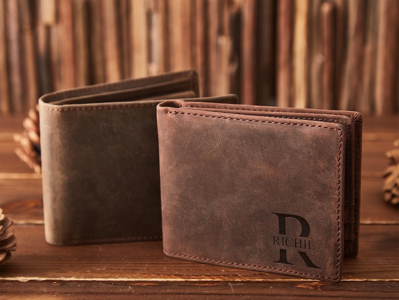 May include: Two brown leather wallets are displayed on a wooden surface. One wallet is open, revealing the interior, while the other is closed and features the name "RICHIE" with a stylized "R" initial. The wallets have a classic, rugged appearance.