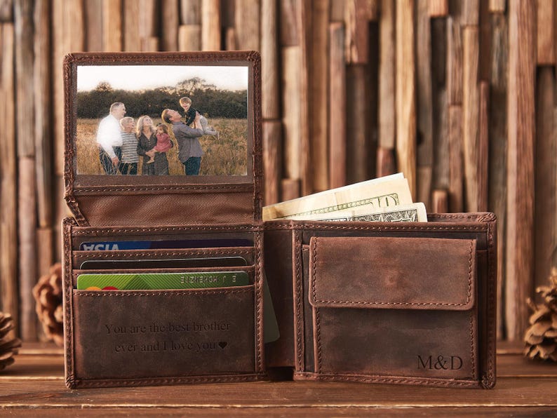 May include: Open brown leather wallet with a family photo, credit card slots, and cash. The wallet is engraved with the text "You are the best brother ever and I love you." and initials "M&D".