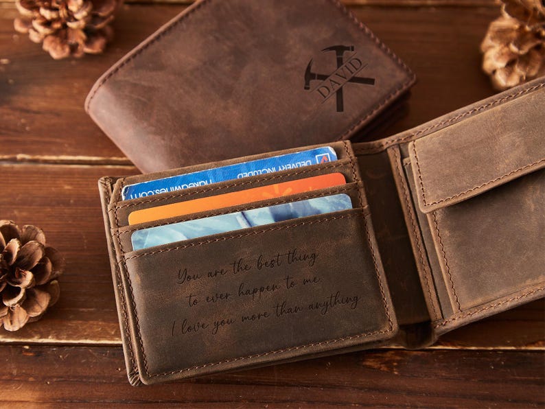 May include: Open brown leather wallet with multiple card slots, displaying credit cards. The wallet is open, revealing a message: "You are the best thing to ever happen to me. I love you more than anything." A second wallet is in the background.