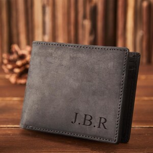May include: A gray leather wallet with the initials "J.B.R" embossed on the front. The wallet has a stitched border and a dark interior. The background features wooden planks and pine cones.
