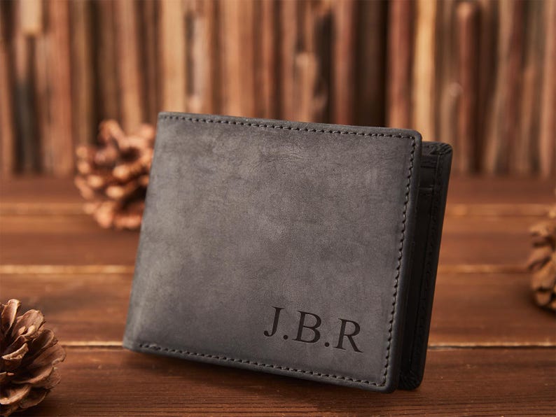 May include: A gray leather wallet with the initials "J.B.R" embossed on the front. The wallet has a stitched border and a dark interior. The background features wooden planks and pine cones.