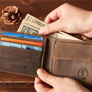 May include: A brown leather wallet with a bi-fold design, holding several credit cards and US dollar bills. The interior of the wallet has the engraved text: "I love you more than all the stars in every universe combined!" A monogram "K" is on the exterior.