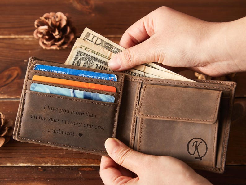 May include: A brown leather wallet with a bi-fold design, holding several credit cards and US dollar bills. The interior of the wallet has the engraved text: "I love you more than all the stars in every universe combined!" A monogram "K" is on the exterior.