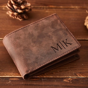 May include: A brown leather wallet with the initials "MK" embossed on the front. The wallet is rectangular and has a stitched border. The background includes pine cones on a wooden surface.